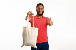 African American Man Showing White Eco Bag And Gesturing Thumbs Up Posing Standing On White Studio Background, Smiling To Camera. I Like Ecology Friendly Lifestyle, No More Plastic Concept photo