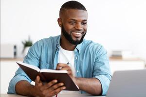 Black Millennial Freelancer Guy Taking Notes To Notepad And Working On Laptop At Home Office, Young African American Man Looking At Computer Screen And Writing In Notepad, Planning Business Schedule photo