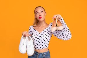 Elegance Or Sport. Funny pensive woman holding pair of high heels shoes and white sneakers in hands, thinking and choosing what to wear, looking up, isolated over yellow background wall, free space photo