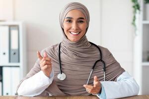 Smiling female doctor young beautiful woman in hijab with stethoscope making consultation in her cabinet, sitting at workdesk with medical chart, notebook and laptop in clinic, copy space photo