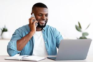 Black Male Entrepreneur Talking On Cellphone And Working On Laptop In Office. Smiling Millennial African American Businessman Sitting At Desk With Computer And Using Mobile Phone, Free Space photo
