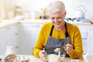 Joyful grandmother wearing apron making dough at kitchen, adding flour, copy space. Happy elderly lady baking pastry cookies or cake at home. Domestic lifestyle, culinary hobby concept photo