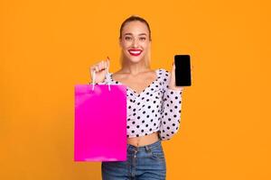 Online Sales And Mobile Application. Smiling woman showing smartphone with black empty screen for mock up template and holding shopping bag, standing and posing at yellow studio wall background photo
