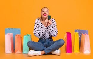 Easy Payment. Portrait of excited stylish young woman holding credit card, ordering purchases and paying online via internet, buying clothes, sitting at yellow studio on the floor with shopping bags photo