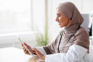 Female doctor muslim woman using digita tablet while working in clinic or hospital, having online appointment with patient, using application, reading medical articles, side view, copy space photo