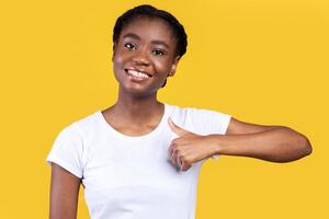 Black Young Lady Gesturing Thumbs Up Smiling To Camera Posing Standing Over Yellow Background In Studio. I Like It, Thumbs-Up Gesture Concept. Woman Showing Thumbs In Approval photo