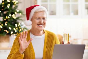 Positive senior woman in Santa hat drinking champagne and having conference with her family, sitting at decorated kitchen and using laptop, waving at camera. Celebrating xmas or New Year photo