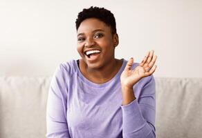 Portrait of positive african american woman smiling, laughing and looking at camera, sitting on sofa at home. Young black female gesturing with hand, touched by compliment, hearing funny joke photo