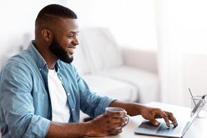 Telecommuting Concept. Portraif Of Young Smiling Black Freelancer Guy Working On Laptop At Home, Positive African Guy Sitting At Desk With Computer, Enjoying Remote Work, Side View With Copy Space photo