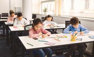 Schooling Concept. Happy Diverse Multiethnic Children Studying In School, Sitting At Tables With Social Distance, Taking Notes And Writing in Copybook. International Students Learning New Material photo