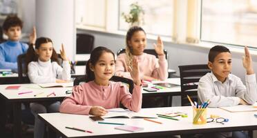 First Day Of School. Multiethnic group of diverse little classmates sitting at tables in modern classroom, raising hands up for an answer, discussing new theme with teacher, participation concept photo