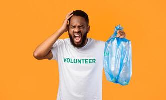 Mad African American Volunteer Man Shouting Holding Plastic Garbage Bag Full Of Litter Expressing Anger And Rage Standing Over Yellow Studio Background. Waste Sorting And Ecology Problems photo
