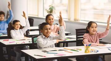 Ready For An Answer. Diverse group of multicultural cheerful primary schoolkids sitting at table in classroom and raising hands, studying and learning. Portrait of positive little elementary students photo