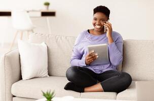 Always In Touch. Happy young plus size african american woman talking on mobile phone, sitting on the couch at home and holding digital tablet. Communication And Technology Concept. photo