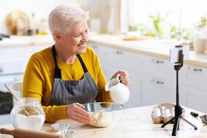 Food, culinary bloggers concept. Smiling senior woman food vlogger wearing apron and broadcasting while cooking, using mobile phone on tripod in kitchen, baking pie or biscuit, copy space photo