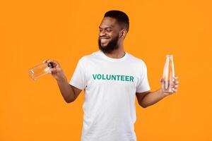 Cheerful Black Male Volunteer Choosing Between Glass And Plastic Bottle Standing Over Yellow Studio Background. No More Plastic, Conscious Consumption Concept photo