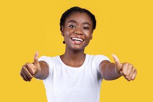 I Like It. Smiling African Woman Gesturing Thumbs Up With Both Hands Smiling To Camera Posing On Yellow Studio Background. Young Black Lady Showing Thumbs Approving And Advertising Something photo