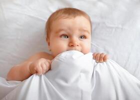 Closeup of adorable blonde baby laying in bed under blanket. Happy little kid infant woke up after sleeping, playing with duvet and looking at copy space, lying in baby bed photo
