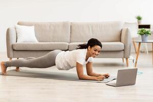 Training At Home. Full length portrait of sporty smiling black woman doing plank while watching online tutorial on laptop computer, exercising in living room on the floor on yoga mat, free space photo