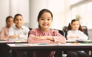 First Day At School. Portrait of smiling diligent asian girl sitting with folded arms at table with group of classmates in blurred background, posing, looking at camera, selective focus. Back To Study photo