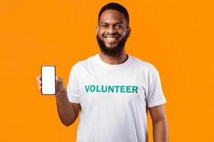 African American Volunteer Man Shows Blank Smartphone Screen Smiling To Camera Standing Over Yellow Studio Background. Great Mobile Application For Volunteering Concept. Mockup photo