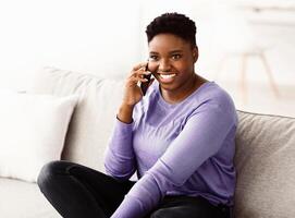 Happy cheerful young black woman talking on the phone at home, smiling lady making call by cellphone sitting on sofa at home, having pleasant or funny conversation speaking by mobile photo