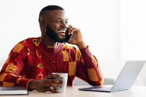 Handsome African Freelancer Guy Talking On Cellphone And Drinking Coffee, Resting At Desk In Home Office, Having Pleasant Phone Conversation, Enjoying Working Remotely And Online Job, Copy Space photo