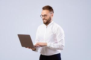 Smiling young guy in office wear using laptop computer over grey studio background. Millennial freelancer working on business project remotely, studying online, having web conference photo