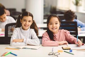 Education And Children Concept. Portrait of happy smiling small girls sitting at desk in classroom at elementary school with group of classmates in background and posing, looking at camera photo