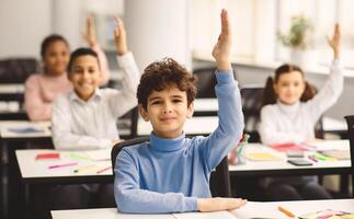 First Day Of School. Portrait of cute boy sitting at table in classroom, group of multicultural small pupils all raising hands up for an answer, studying in modern classroom, selective focus photo