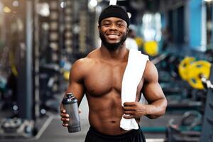 Rest after workout. Happy african american bodybuilder holding water or protein and towel at gym, empty space. Smiling black muscular man having break while exercising at modern gym photo