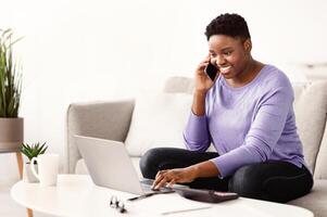 Multitasking At Home. Smiling young plus size black lady making call using her smartphone, sitting on sofa, typing on laptop keyboard. Positive woman talking with client or colleagues about work photo