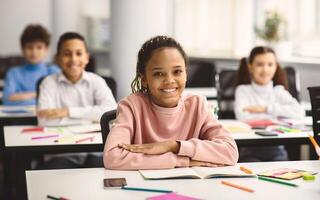 Elementary Education. Portrait of happy small african american girl sitting at table in classroom at primary school with group of classmates and posing, looking at camera and smiling photo