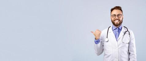 Positive Caucasian male doctor in lab coat showing thumb up gesture and pointing at empty space over grey studio background, banner design. Healthcare and medical concept photo