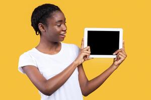 African American Woman Showing Digital Tablet With Blank Computer Screen Advertising Website Or Application Posing Standing Over Yellow Background In Studio. Modern Lifestyle, Gadgets And Technology photo