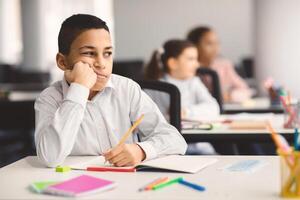 Education And Learning Concept. Portrait of tired and bored small boy sitting at desk in classroom at school, writing in notebook and thinking, looking away at window, resting head on hand photo