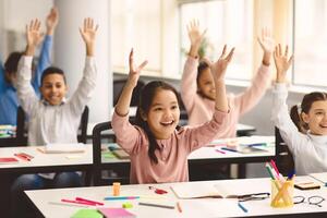 Portrait of multiciltural excited diverse group of small happy elementary school children sitting at desks in classroom, raising hands high up and waving, having fun, learning with pleasure photo