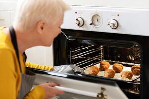 Over shoulder shot of senior grey-haired woman with short hair checking beautiful croissants in oven, closeup. Smiling elderly lady baking at home, sitting next to oven in kitchen, taking pastry out photo