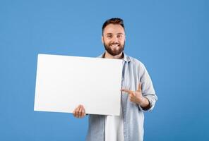 Free space for advertising. Happy Caucasian man pointing at empty white board on blue studio background, mockup for design. Millennial bearded guy with blank billboard template photo