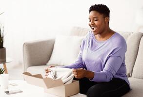 Happy Customer Concept. Portrait of excited curvy black lady opening and unpacking cardboard box parcel, sitting on the couch in living room at home. Young happy female satisfied with delivery service photo