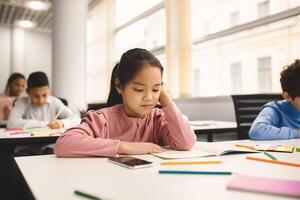 Portrait of focused small asian girl sitting at table in classroom at primary school or kindergarten, reading task in notebook. Reopening and return back to school after coronavirus quarantine photo