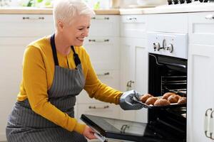 Smiling elderly woman baking pastry at home, taking delicious croissants out of oven, copy space. Joyful senior lady in apron opening oven, checking her pastry, cozy kitchen interior, closeup photo