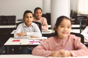 Elementary Education. Portrait of small smiling boy sitting at table in classroom at primary school or kindergarten with group of classmates. Return back to school after covid quarantine photo