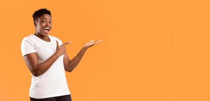 Joyful Plus Size African Lady Pointing Finger Holding Invisible Object Showing And Advertising Your Product Posing Over Yellow Background, Smiling To Camera. Studio Shot, Free Space photo