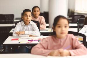 First Day At School. Portrait of focused diligent boy sitting with folded arms at table with group of classmates in blurred background, listening to teacher, selective focus. Back To Study photo