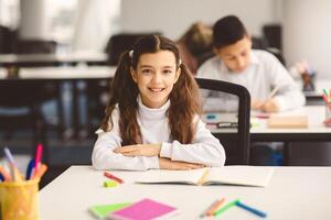 First Day At School. Portrait of smiling diligent small girl with ponytails sitting at table in classroom at elementary school with group of classmates in background and posing, looking at camera photo