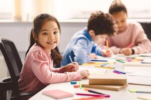 Academic Concept. Smiling junior asian school girl sitting at desk in classroom, writing in notebook, posing and looking at camera. Group of diverse classmates studying in the background photo