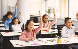 Return Back To School. Multiciltural diverse group of happy small elementary schoolchildren sitting at desks in classroom, raising hands up for an answer, learning with pleasure. Social distance photo