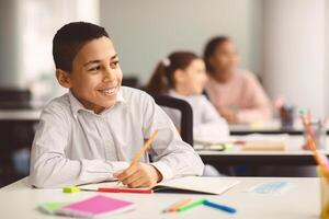 Elementary And Primary School Education. Portrait of smiling small boy sitting at table in classroom, writing or drawing in notebook, looking away at window. Reopening and return back to school photo