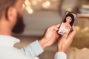 Long Distance Relationship. Back over the shoulder view of black man having online virtual date with his surprised girlfriend, holding and using smartphone app for videochat. Remote Dating photo